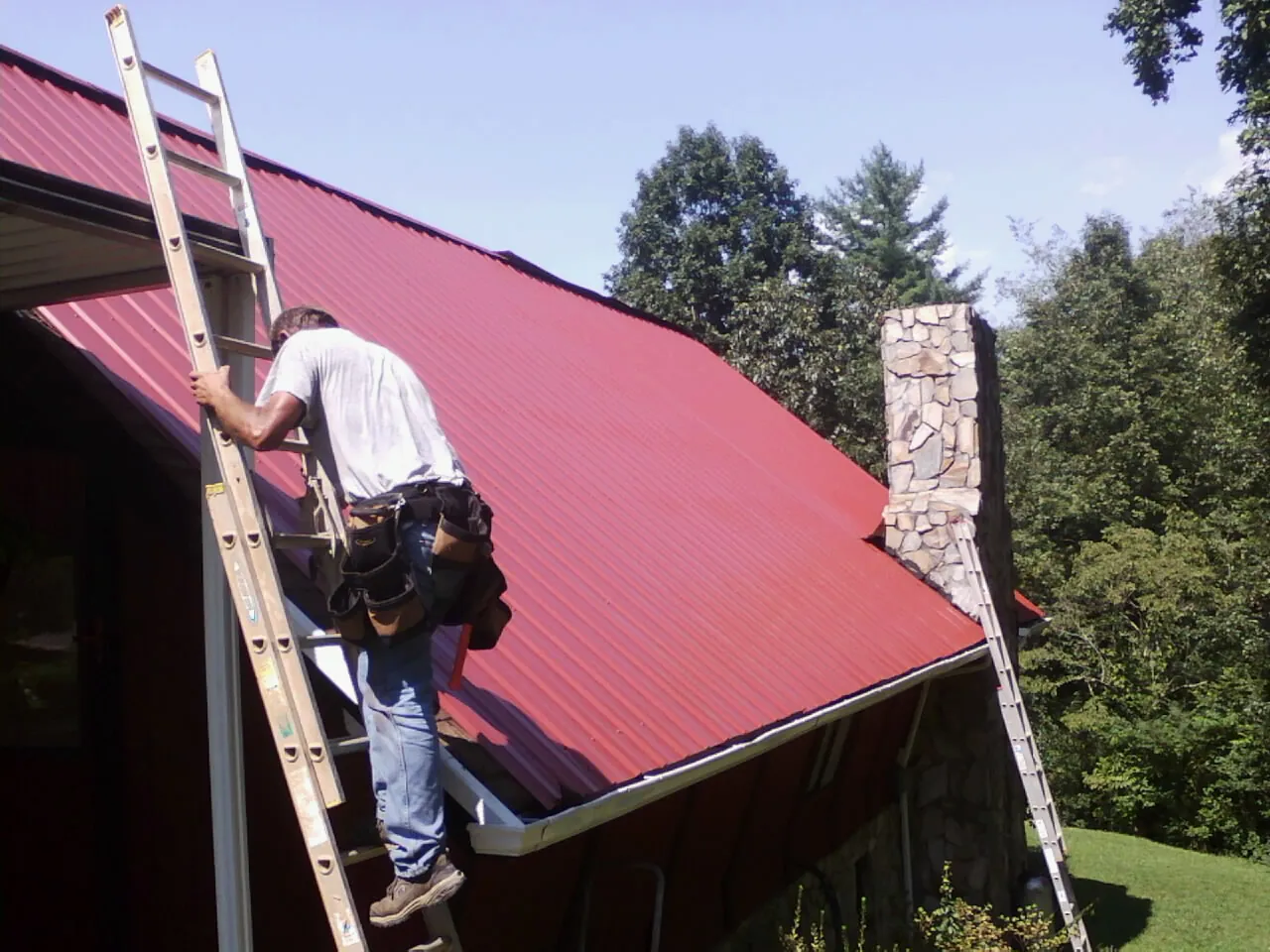 Roofer on a ladder beside a red metal roof and stone chimney — Soffit & Fascia Repair by Roofing Brothers in Galva